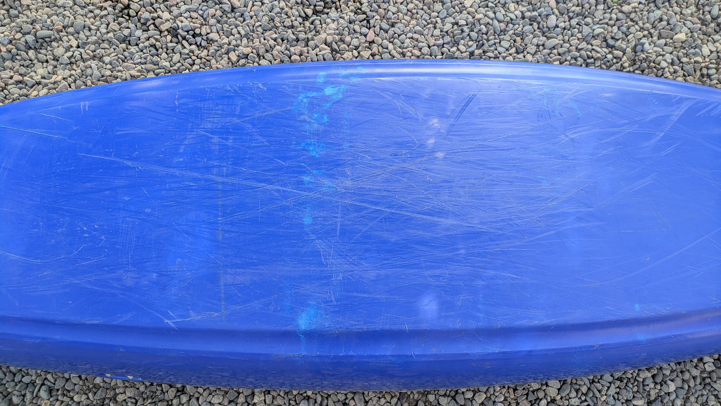 A scratched blue surfboard rests on small, round pebbles beside a used Dagger Demo Rewind LG kayak.