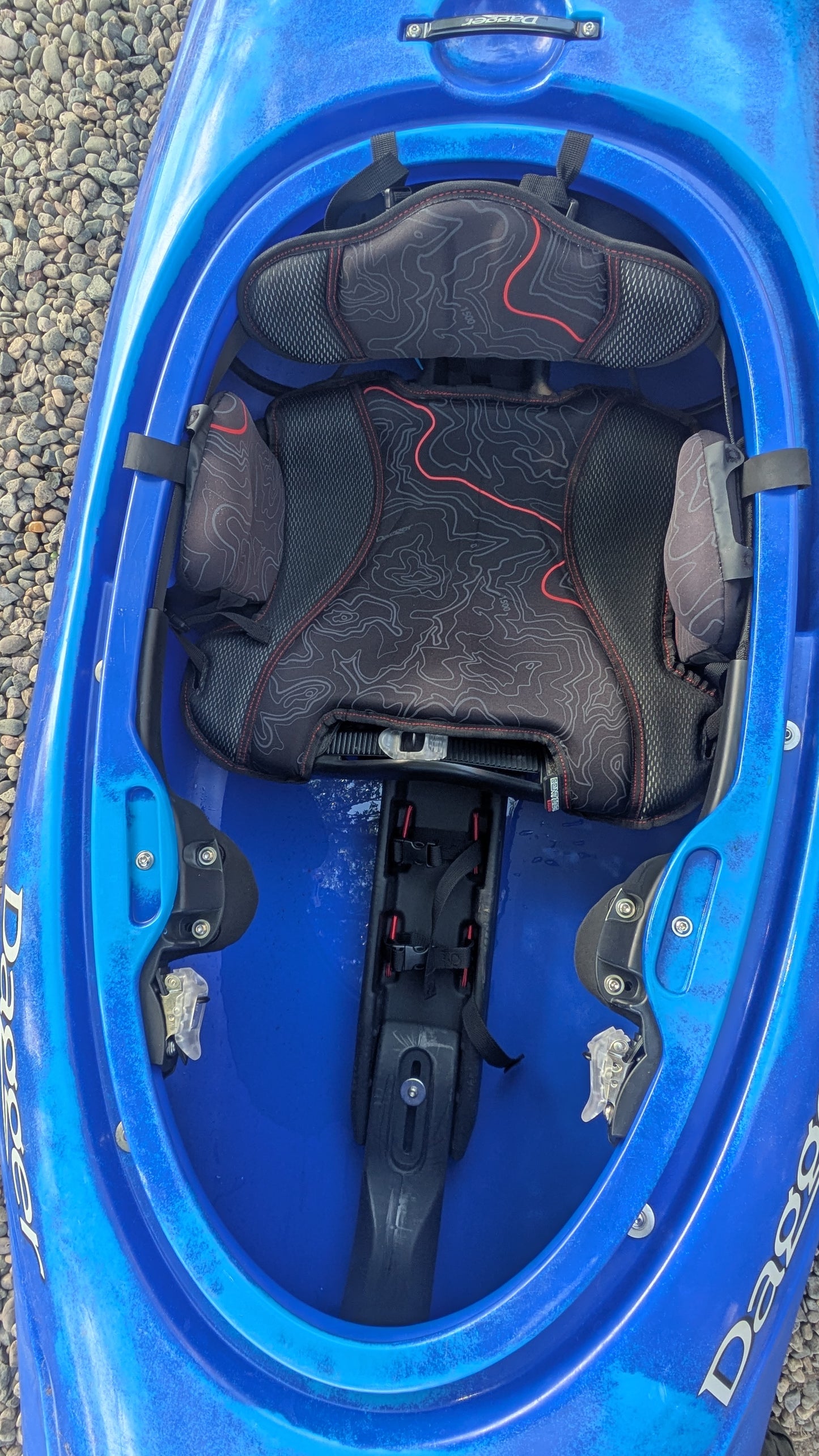 Top view of the interior of a blue Demo Rewind LG kayak by Dagger, showing padded seat, foot braces, and cockpit rim, with a few pebbles visible outside.