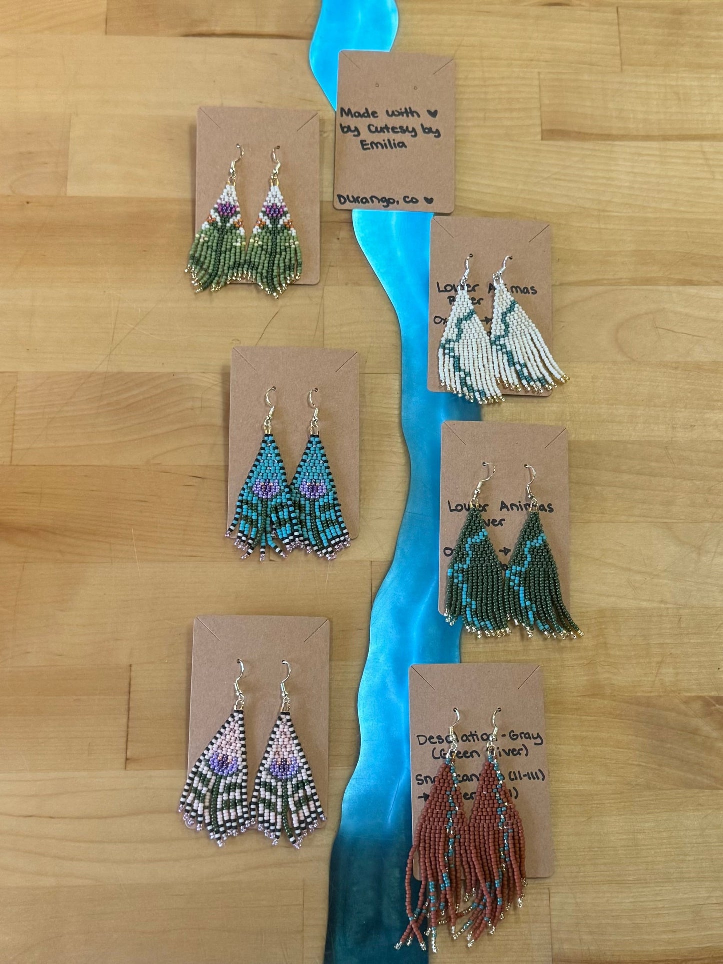 Cutesy By Emilia Earrings by 4CRS are showcased on a wooden surface, with each handcrafted beaded pair attached to labeled cards detailing their design and origin. Some pairs feature exquisite desert flower designs, adding a unique touch to this charming collection.