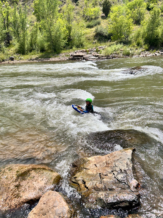 Wearing a green helmet, a kayaker grips a bodyboard and navigates rocky river rapids, capturing the thrill of adventure in 4CRS Paddle School’s Send-It Summer Finale.