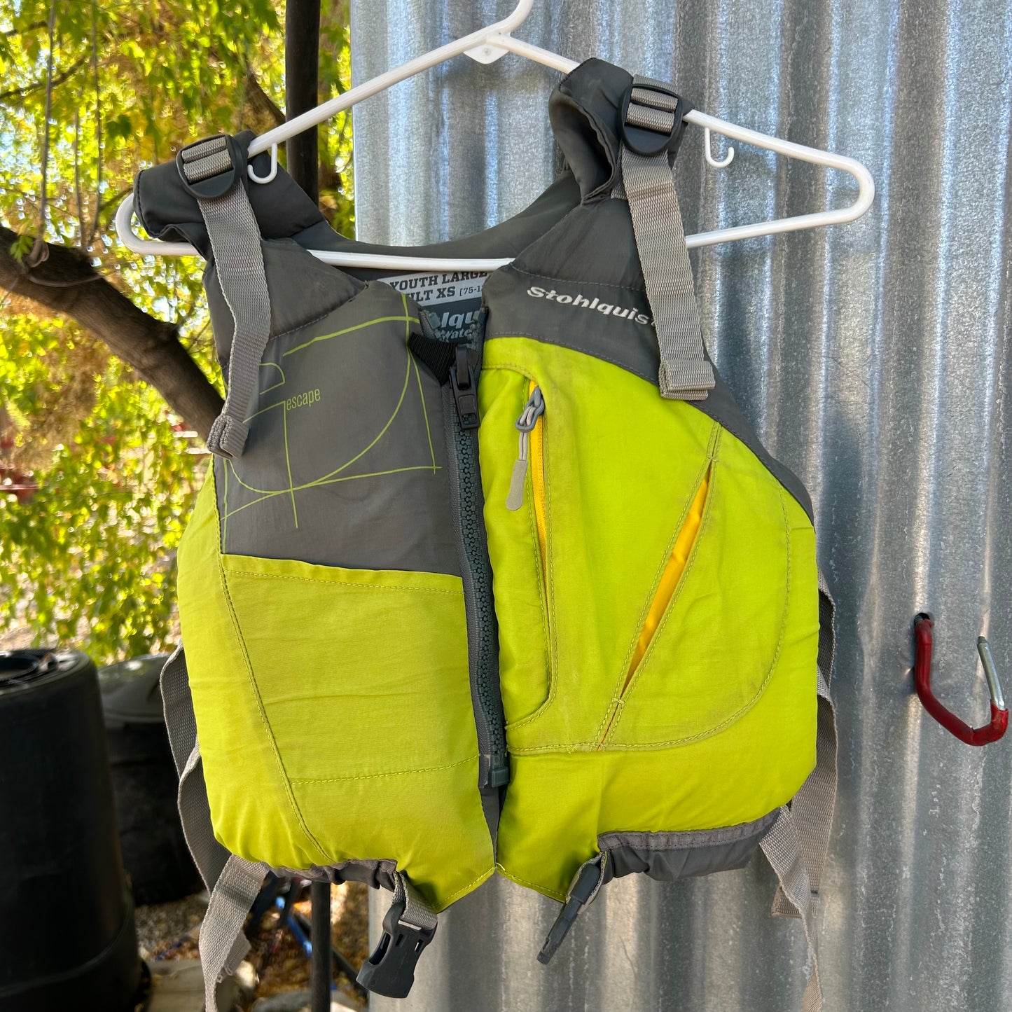 A "Stohlquist" branded Demo Escape Youth/Adult XS PFD in green and gray hangs on a white hanger against a corrugated metal backdrop, showcasing its sleek design and functionality.