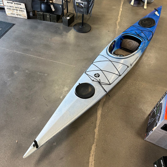 A Consignment Perception Carolina 14.5 touring kayak by Perception, in blue and gray, is shown indoors on a concrete floor with two storage hatches and deck bungee cords.
