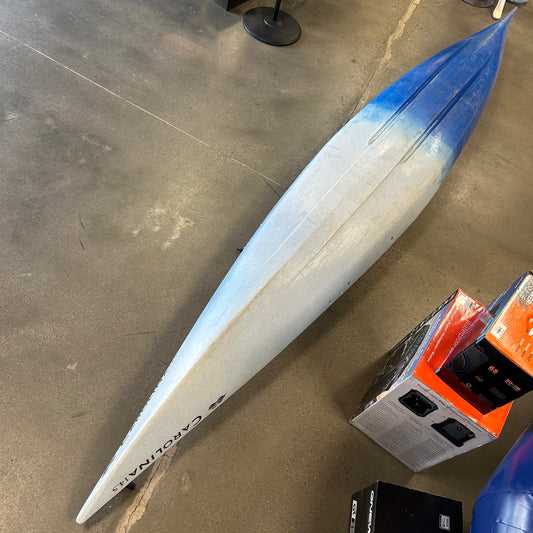 A Consignment Perception Carolina 14.5 touring kayak by Perception, in blue and white, is lying upside down on a concrete floor beside some boxes.