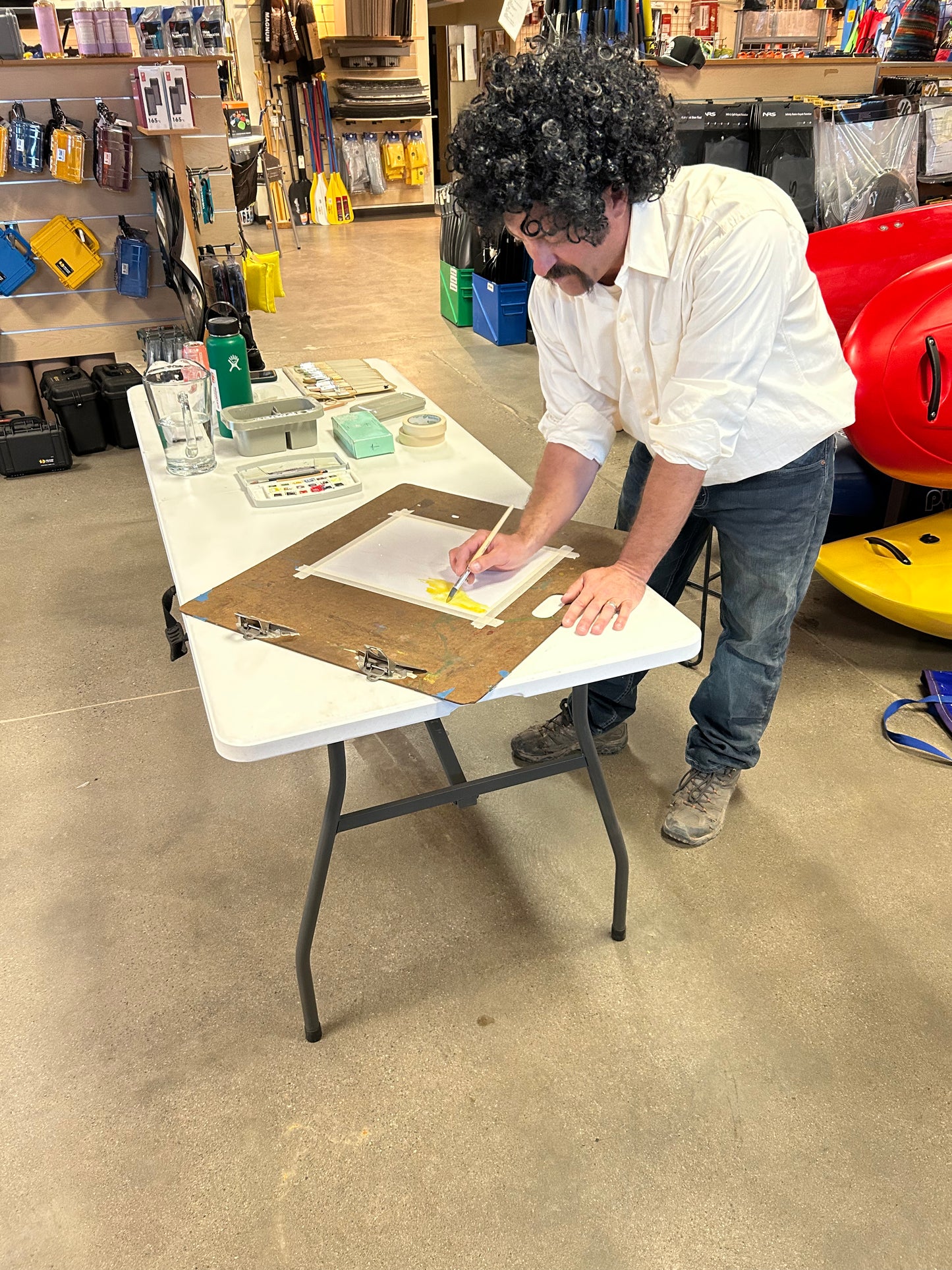 An artist joins the Paint and Sip - Fall River Scene class by 4CRS Paddle School, painting on canvas at a folding table inside a store stocked with outdoor gear.