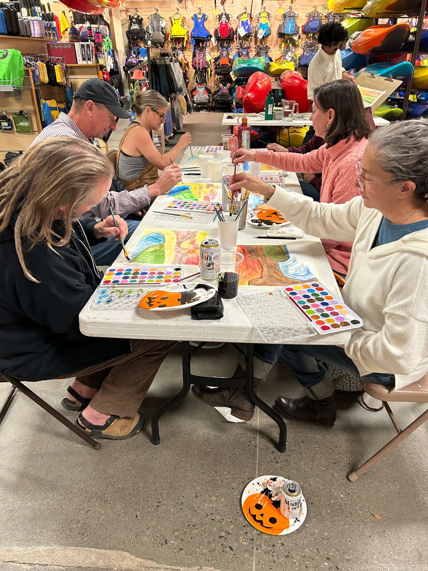 Five adults enjoy the Paint and Sip - Fall River Scene class by 4CRS Paddle School, painting with art supplies at a festive Halloween-themed table. Kayaks and outdoor gear are visible in the store background.