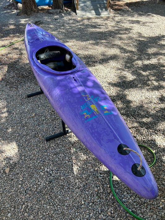 A Used Prijon Canyon kayak from 4Corners Riversports in purple HTP plastic, visibly worn, sits on a stand over gravel outdoors; a green garden hose lies nearby and tree shadows are cast across the scene.