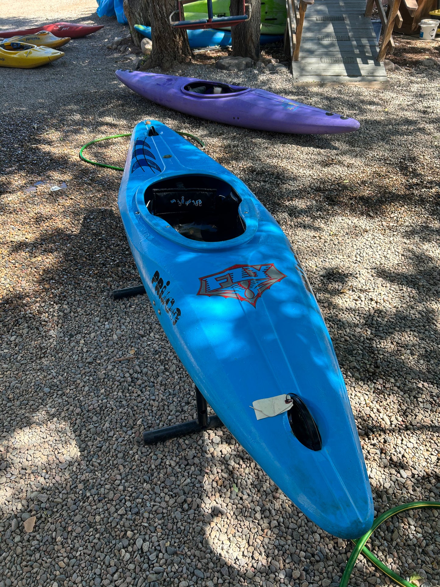A blue Used Prijon Fly kayak from 4Corners Riversports with a planing hull sits on a metal stand over gravel, with a purple playboat and other kayaks visible nearby among trees.