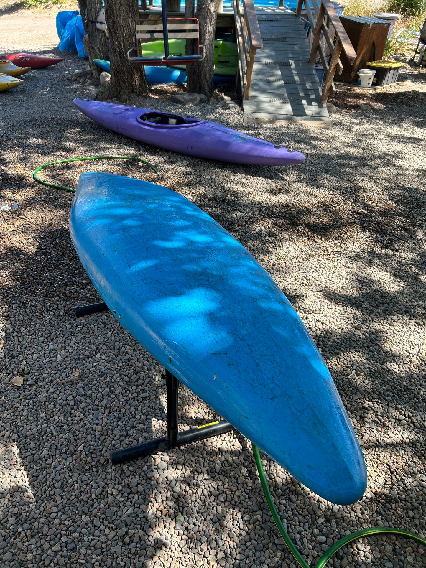 A blue Used Prijon Fly from 4Corners Riversports sits upside down on a metal stand, with a purple kayak and other gear nearby on gravel, close to trees and a wooden ramp.