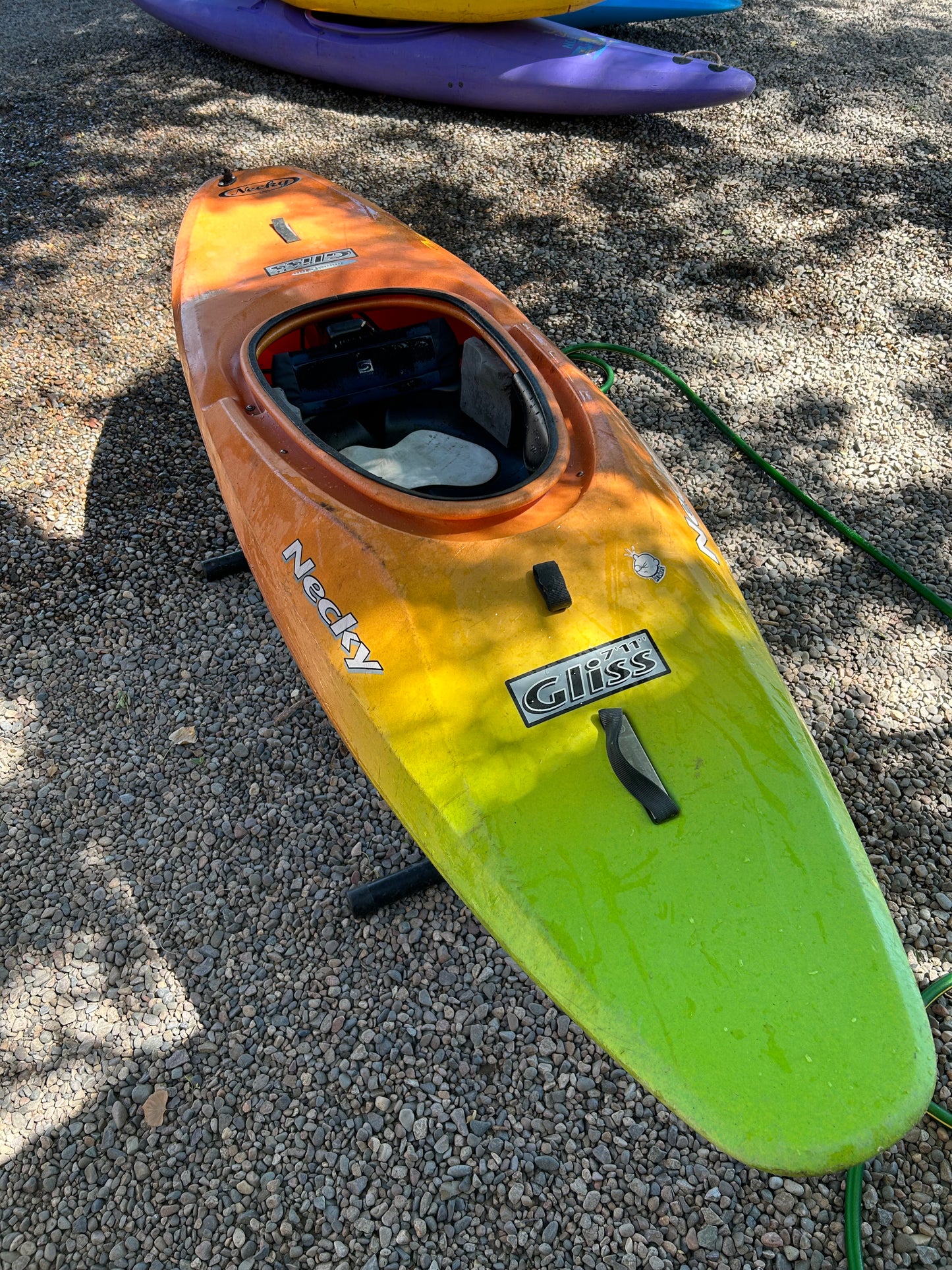 A Used Necky Gliss kayak from 4Corners Riversports in green and orange rests on gravel, with a purple used kayak partly visible behind it.