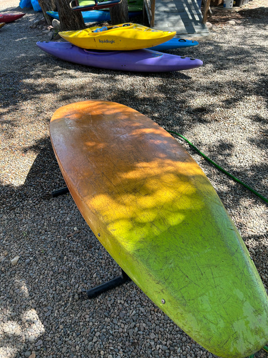 A yellow and green paddleboard rests on a stand on gravel, with two kayaks—a yellow used kayak and a purple Used Necky Gliss from 4Corners Riversports—in the background.