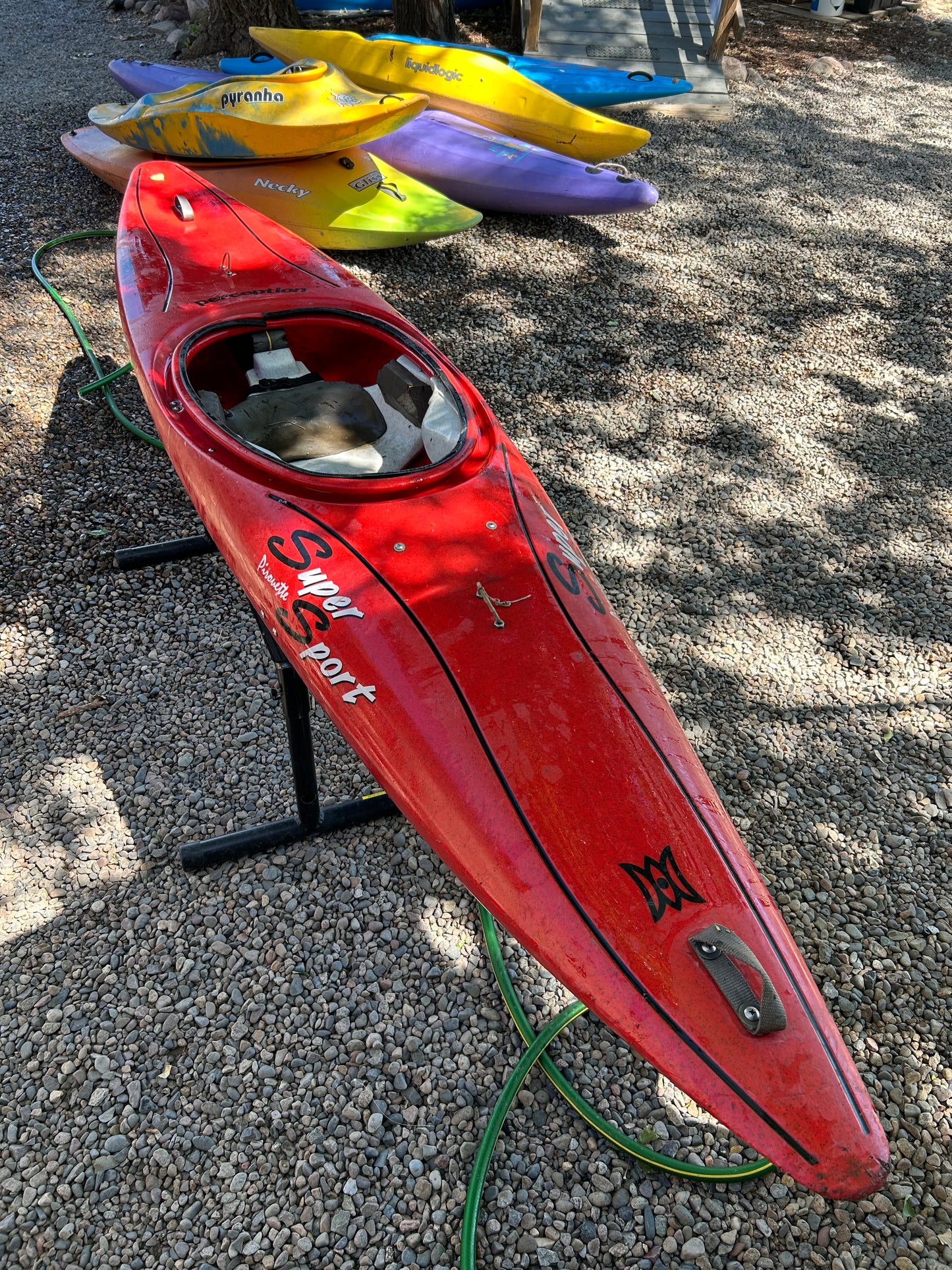 A used Perception Super Sport kayak from 4Corners Riversports, bright red with visible text, is displayed outdoors on stands among other colorful kayaks. Nearby, a green hose rests on the gravel surface.