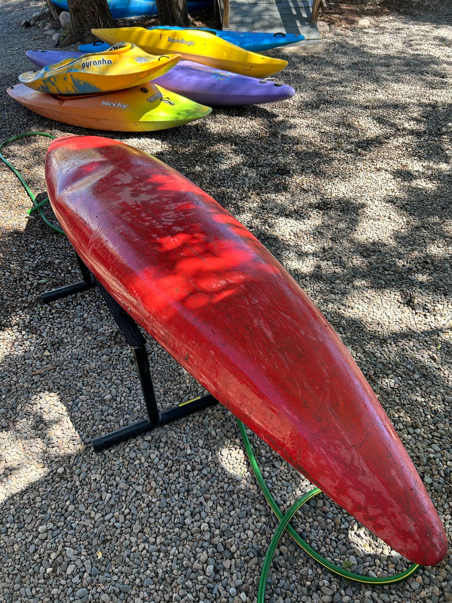 A used Perception Super Sport kayak from 4Corners Riversports is resting upside down on a stand, with several other colorful kayaks lying on the gravel in the background.