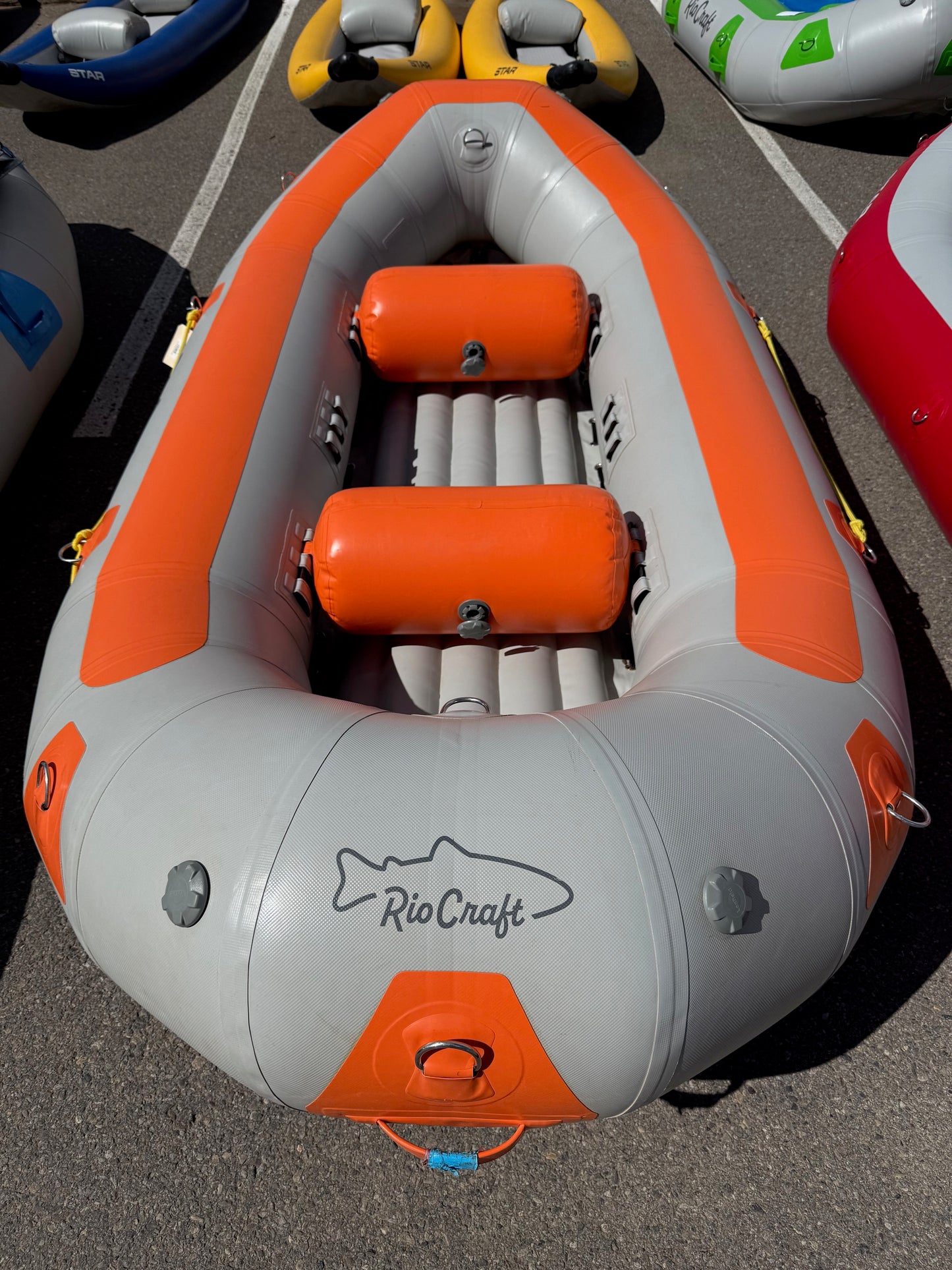 A gray and orange Rio Craft 10.5' Demo inflatable raft with two orange seats is shown on asphalt among other rafts.