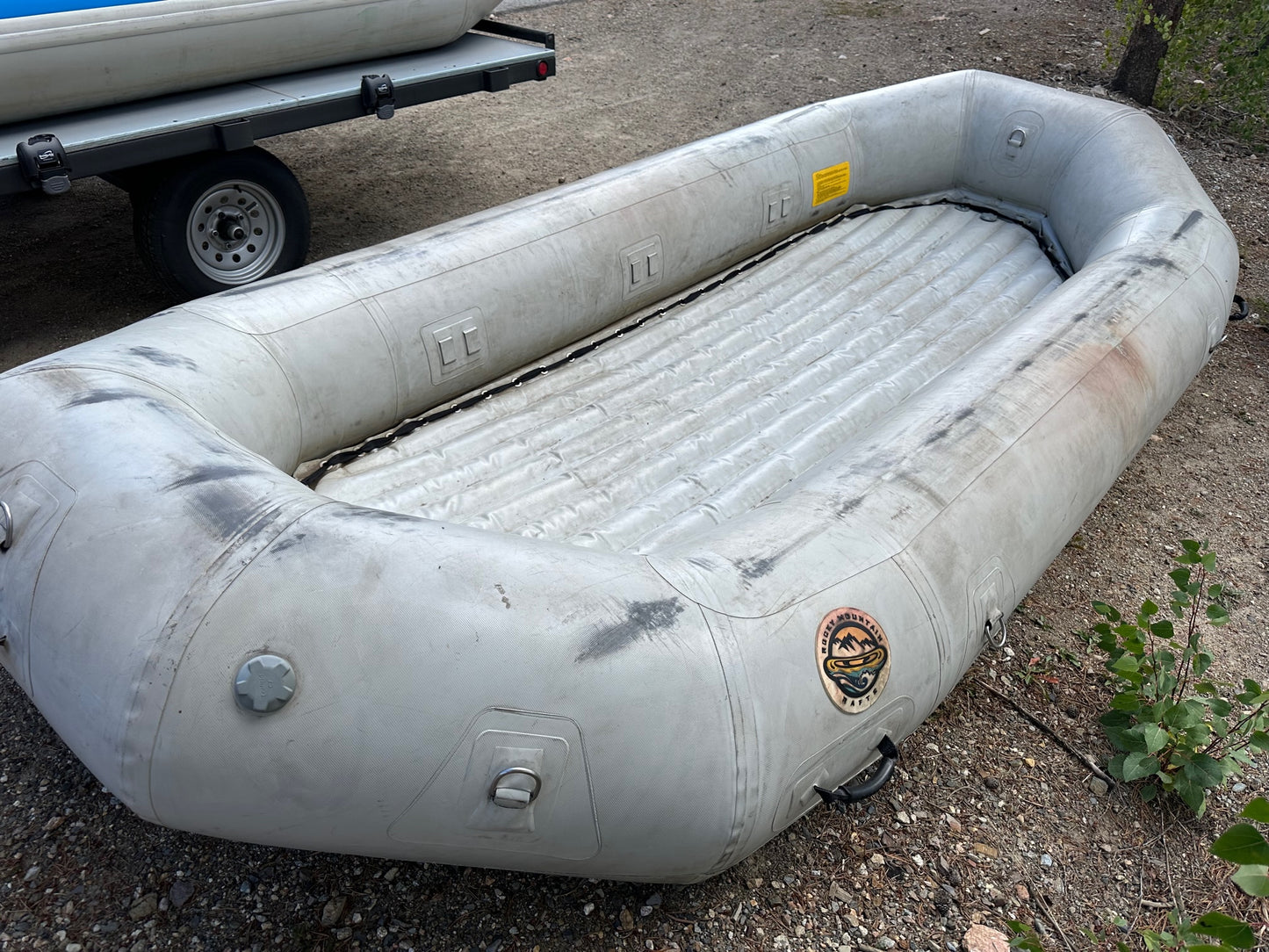 A fair condition Used 14' Rocky Mountain Raft, self-bailing inflatable raft sitting on the ground next to a trailer. (Brand Name: 4Corners Riversports)