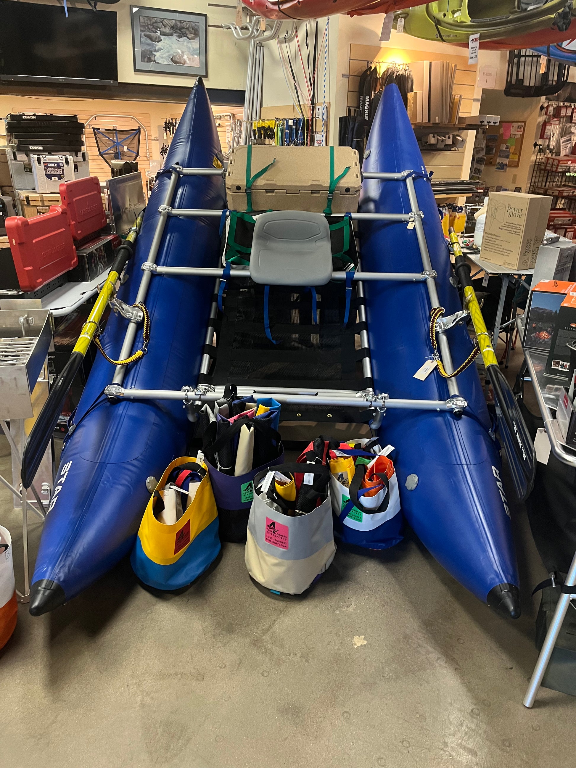 The 13' Star Kima Package by NRS, a blue inflatable cataraft with a central seat and Cataract SGG oars, is displayed indoors amid colorful gear bags and store equipment.