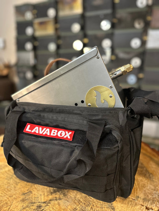 The Hekla Portable Campfire / Stove Combo by LavaBox, with a brass fitting and logo, is partially inside a black LavaBox bag on a wooden surface with shelves in the background.