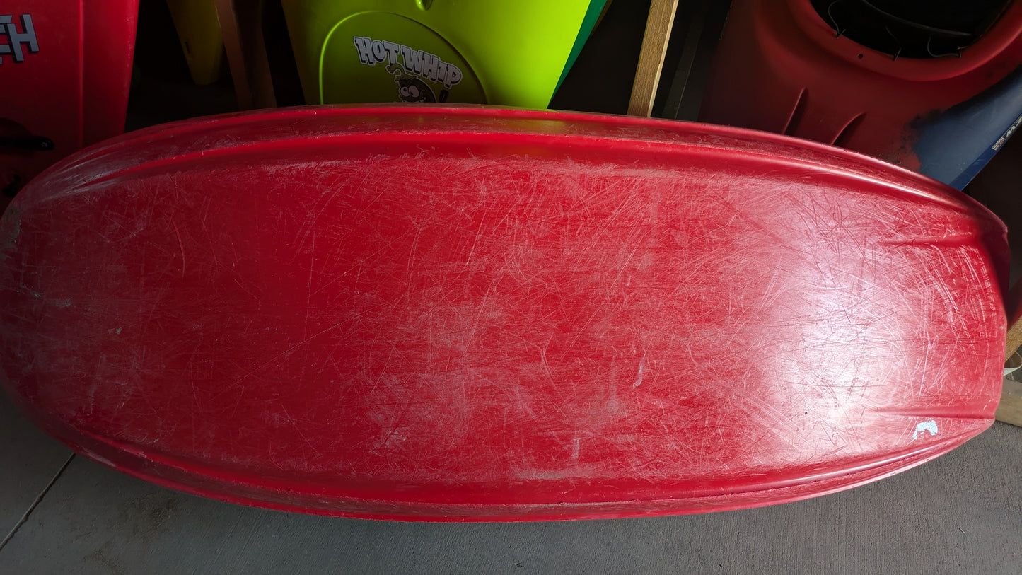 A Jackson Kayak Demo Rockstar V MD with noticeable scratches rests upside down on a concrete floor; colorful sleds, another used kayak, and various objects are partially visible in the background.