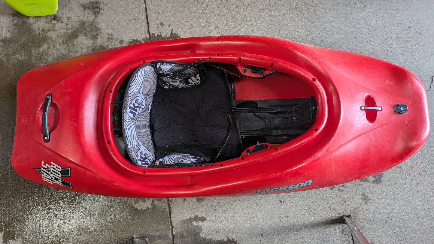 A Jackson Kayak Demo Rockstar V SM in red rests on a concrete surface, seen from above with its seat, footrests, and cockpit interior visible.