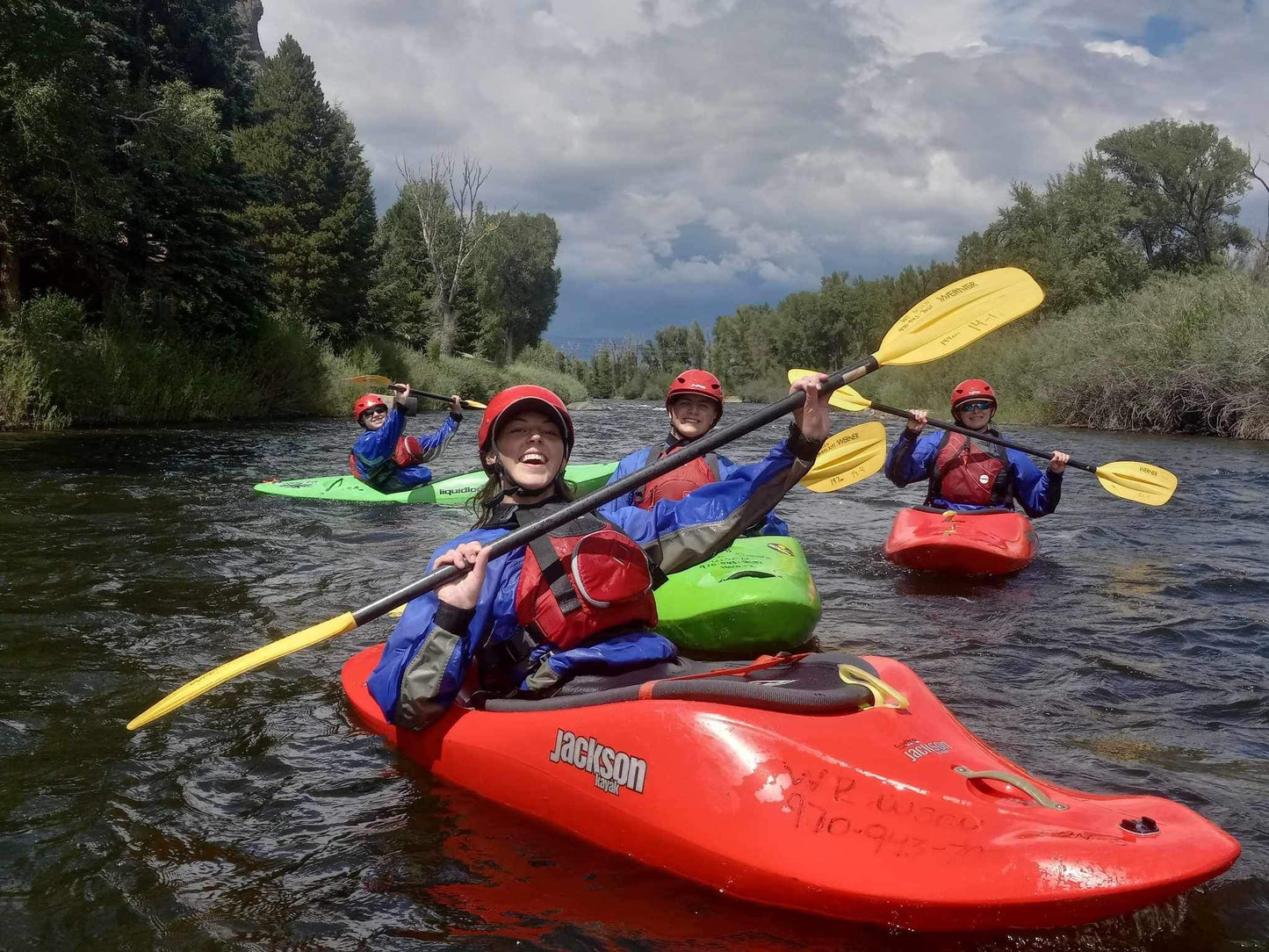 Four adventurers in helmets and life jackets relish "Pedal / Paddle with Devo!" by 4CRS, kayaking through the tree-lined river in vibrant kayaks. The cloudy sky enhances this thrilling summer camp experience.