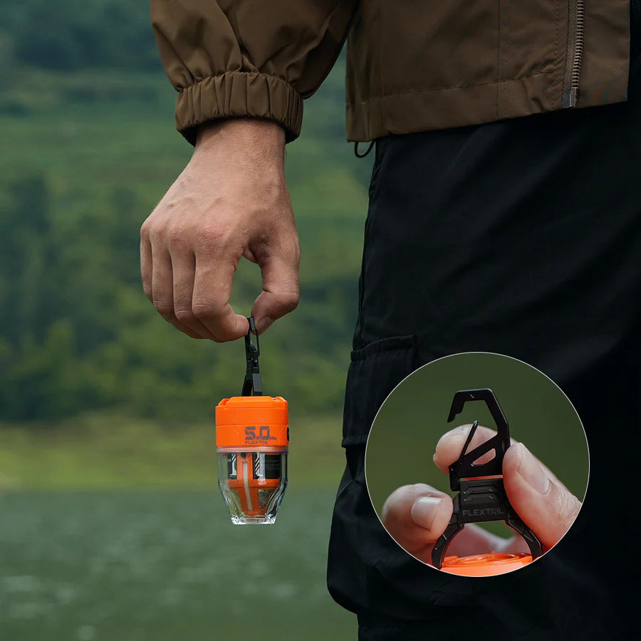 A person holds a compact orange Flextail MAX PUMP 3 camping lantern by a black hook; close-up inset shows the "Flextail" brand and MAX PUMP 3 label. Perfect for packraft trips thanks to powerful 5kPa pressure, pictured outdoors near water and greenery.