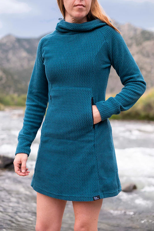A woman stands outdoors by a river, wearing the River Station Gear Z - Fleece Hooded Dress, crafted from recycled polyester Z-Pattern fabric with a high collar and front pocket. Mountains and a cloudy sky complete the serene backdrop.