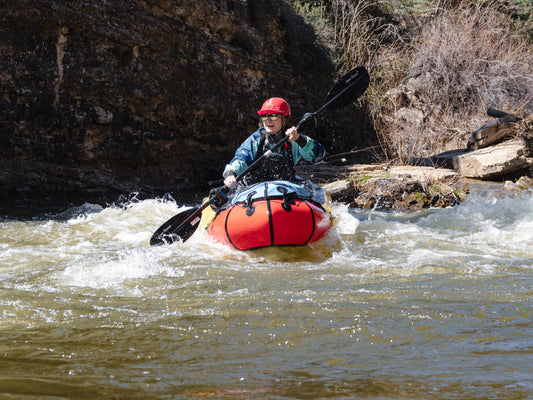 A kayaker wearing a helmet and sunglasses navigates whitewater rapids near a rocky riverbank in Durango, CO—ideal for those seeking the ACA Packraft Instructor Certification from 4CRS Paddle School.