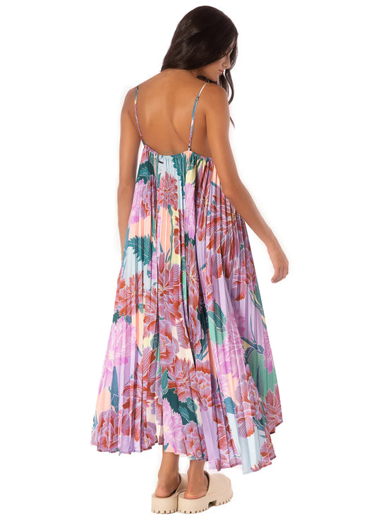 A woman with long dark hair faces away, wearing the Maaji Parrot Dress—a colorful floral maxi with thin straps and an open back. She completes her look with chunky beige sandals.