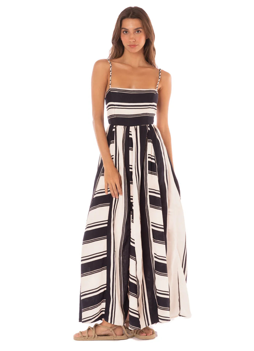 A person stands wearing the Maaji Stripes Ela Long Dress, a sleeveless black and white striped design, paired with sandals against a plain background.