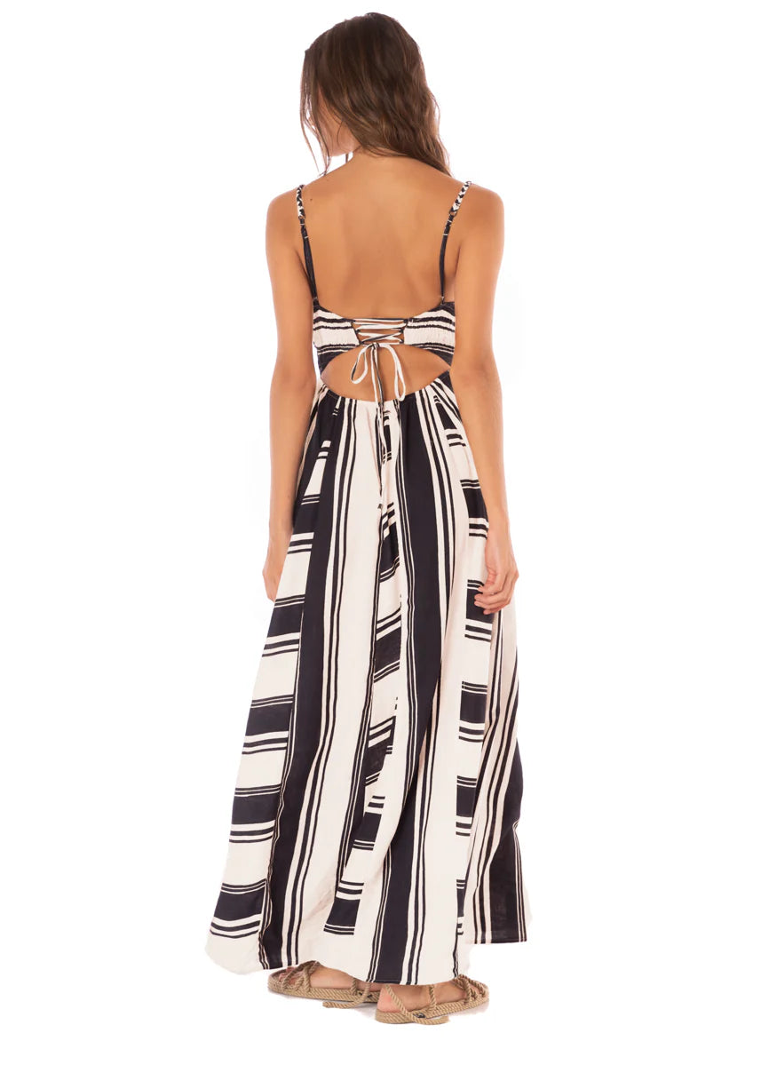 A woman is seen from behind wearing the Maaji Stripes Ela Long Dress, featuring black and white stripes with a back cutout and tie detail, paired with sandals.