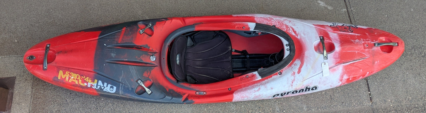 A Used Pyranha Machno whitewater kayak from 4Corners Riversports, featuring a red and black design, rests on a concrete surface as seen from above.