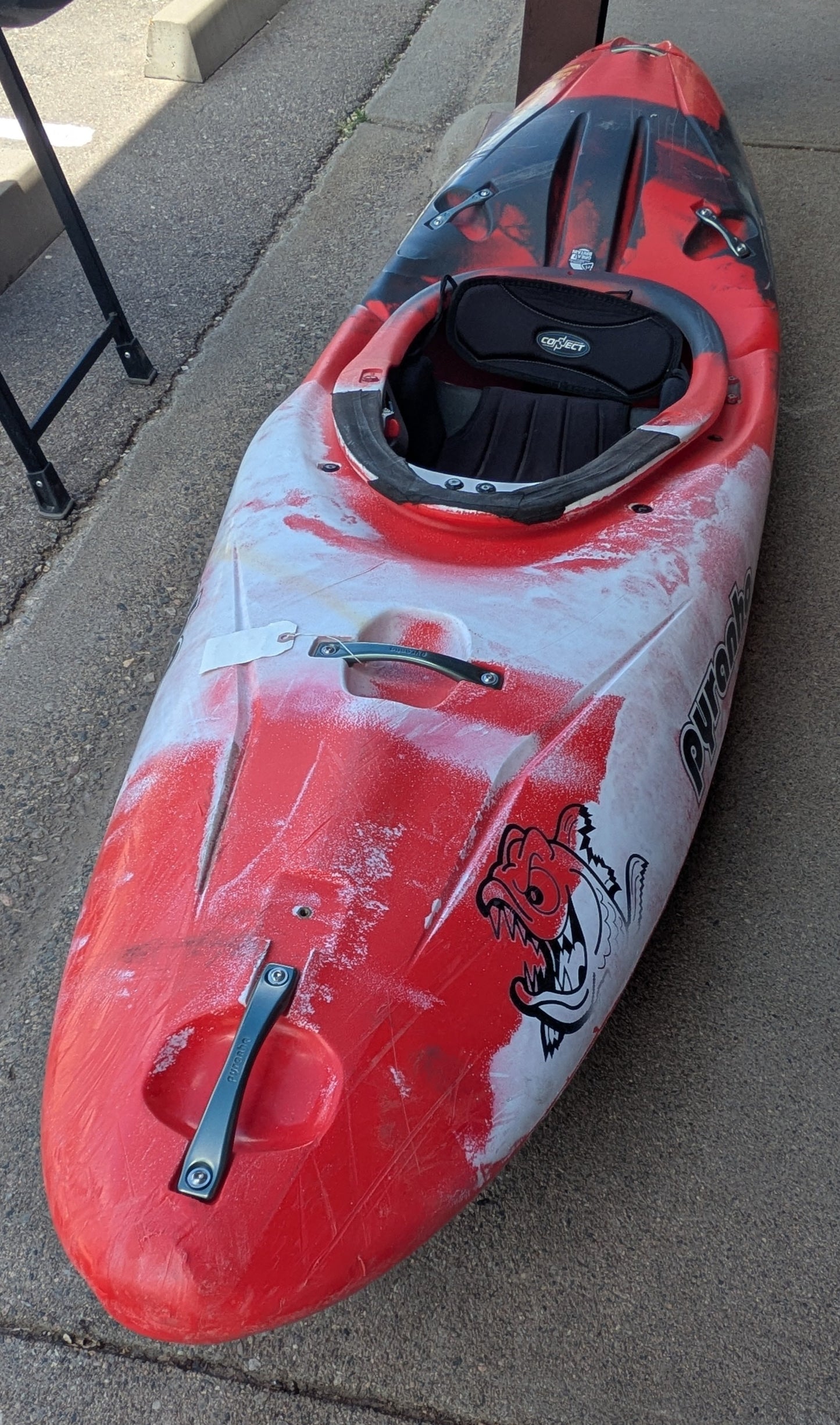 A Used Pyranha Machno whitewater kayak from 4Corners Riversports, featuring a red and white design, seat, handles, and shark-like graphic on the side, is shown resting on a concrete surface.