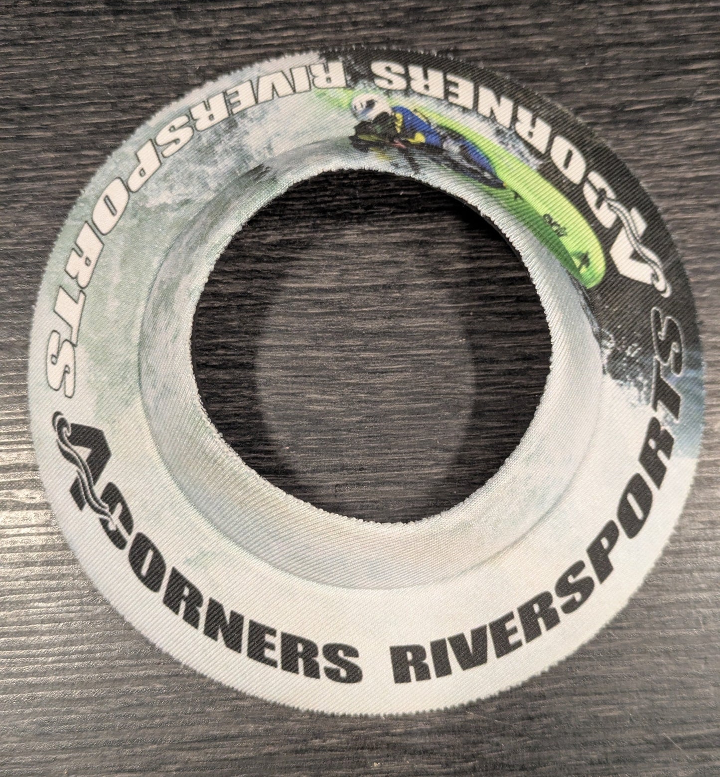 The Lil' Sucker Boozie by 4CRS is a round white fabric band with "4Corners Riversports" text and a kayaker image, designed to securely hold your beer. Shown on a dark wood surface.