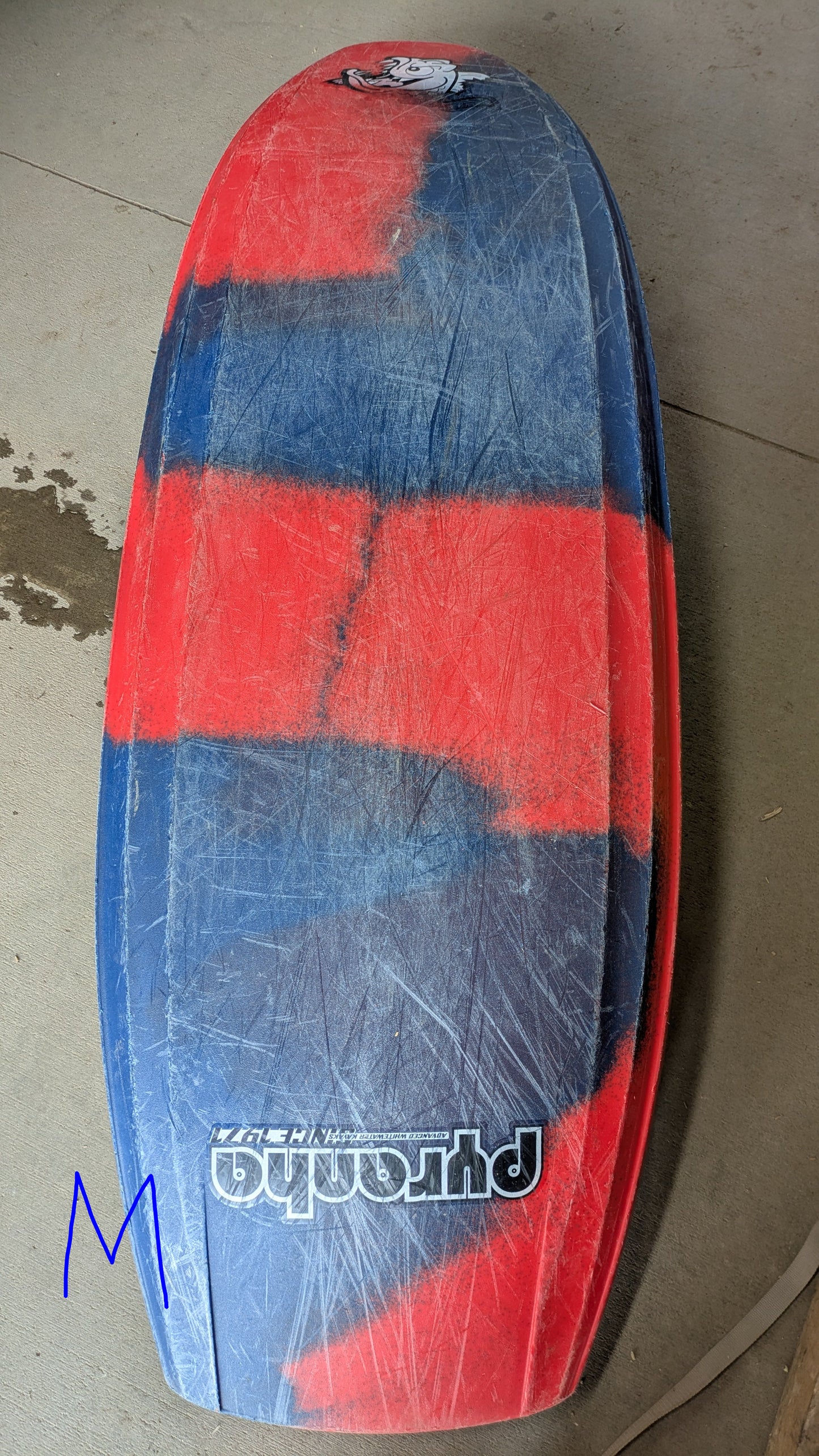 A worn surfboard with red and blue stripes, scratches, and a “Phantom” logo lies on concrete near a blue "M"—much like the Pyranha Demo Helectron used kayak from the demo fleet.