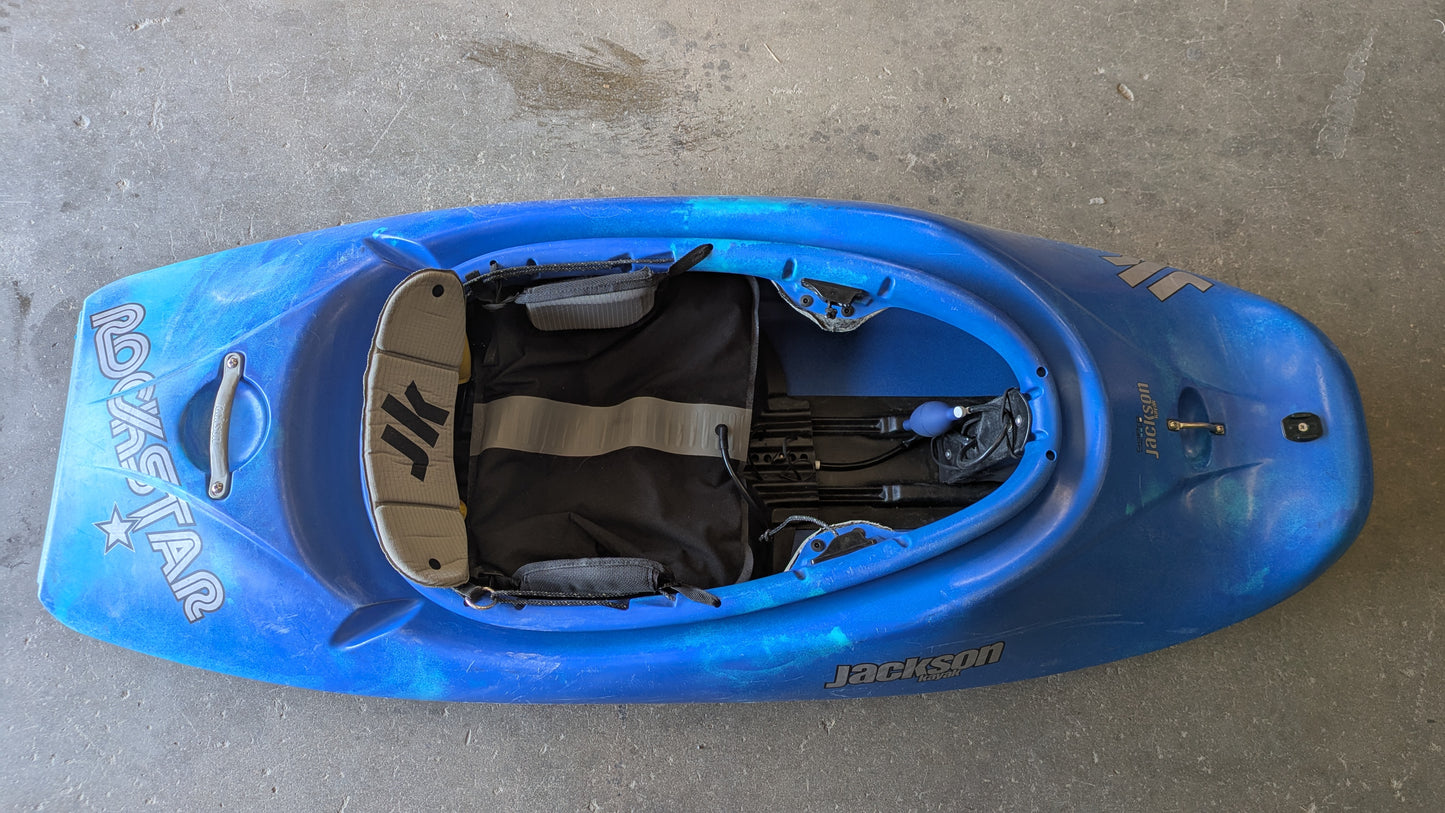 A Consignment Medium Jackson Rockstar 3.0 freestyle kayak from 4Corners Riversports sits on a concrete floor, viewed from above, highlighting its cockpit and seat details.