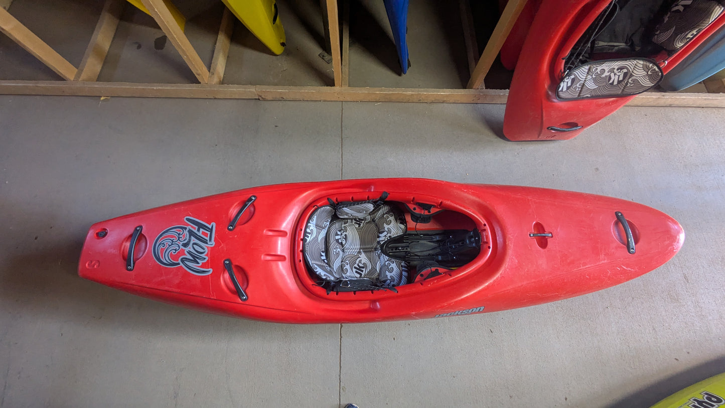 A Jackson Kayak Demo Flow SM in red with black and white interior padding is on a concrete floor, while other kayaks are stored vertically in the background.