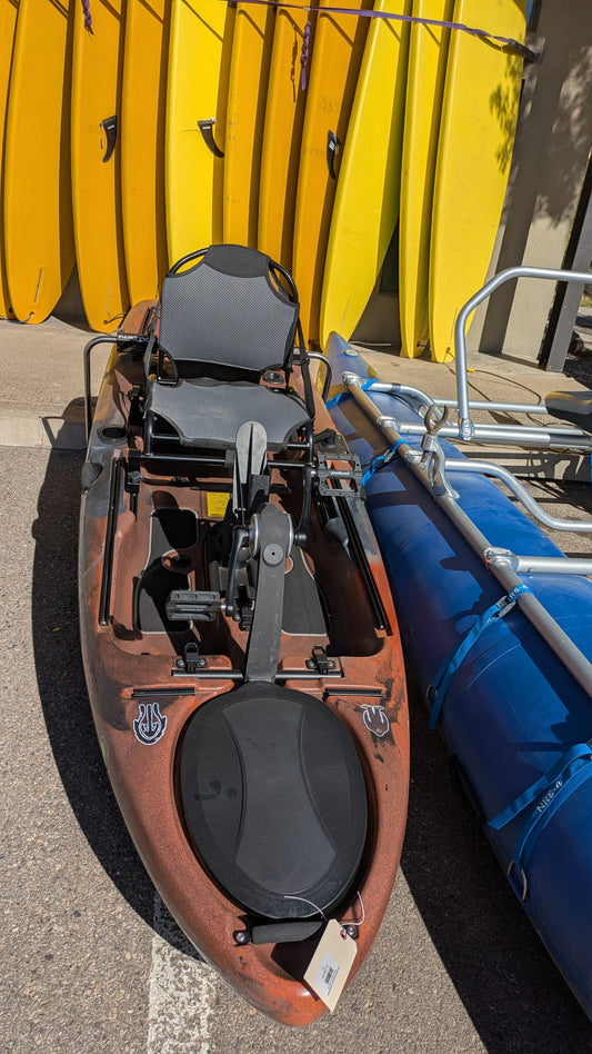 A used Consignment Native Slayer Propel 10 fishing kayak from 4Corners Riversports, featuring a pedal drive and mesh seat, sits in front of yellow paddleboards and beside a blue and silver watercraft.