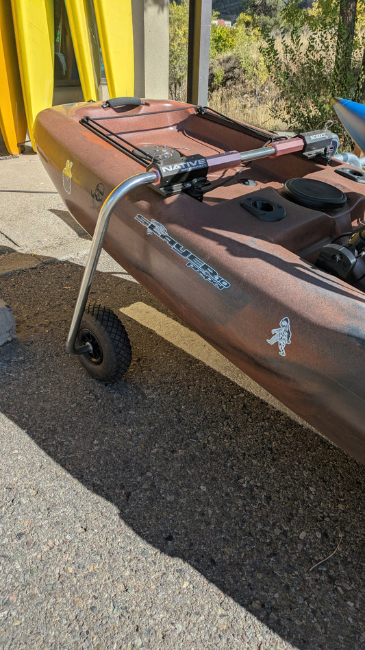 A Consignment Native Slayer Propel 10 fishing kayak from 4Corners Riversports, brown with a metal cart and wheels, rests on pavement near yellow kayaks and trees.