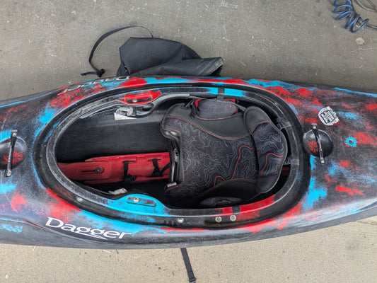 Top view of a Consignment Rewind LG Cosmo whitewater kayak by 4Corners Riversports in black, red, and blue, showing the open cockpit with padded Contour Ergo seat and footrest on a concrete surface.