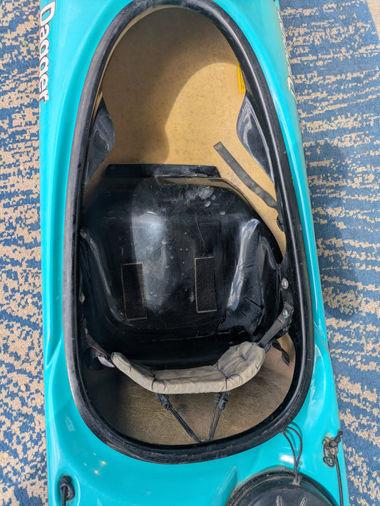 Top-down view of the cockpit on a blue Dagger Sitka Touring/Sea Kayak by Dagger, featuring the seat, backrest, interior fittings, and integrated rudder system on a patterned carpeted floor.