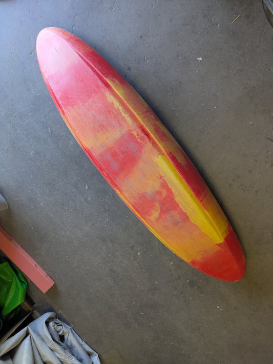 A Used Jackson Zen by Jackson Kayak sits on a concrete floor, its bold red and yellow colors making it stand out among various other items.