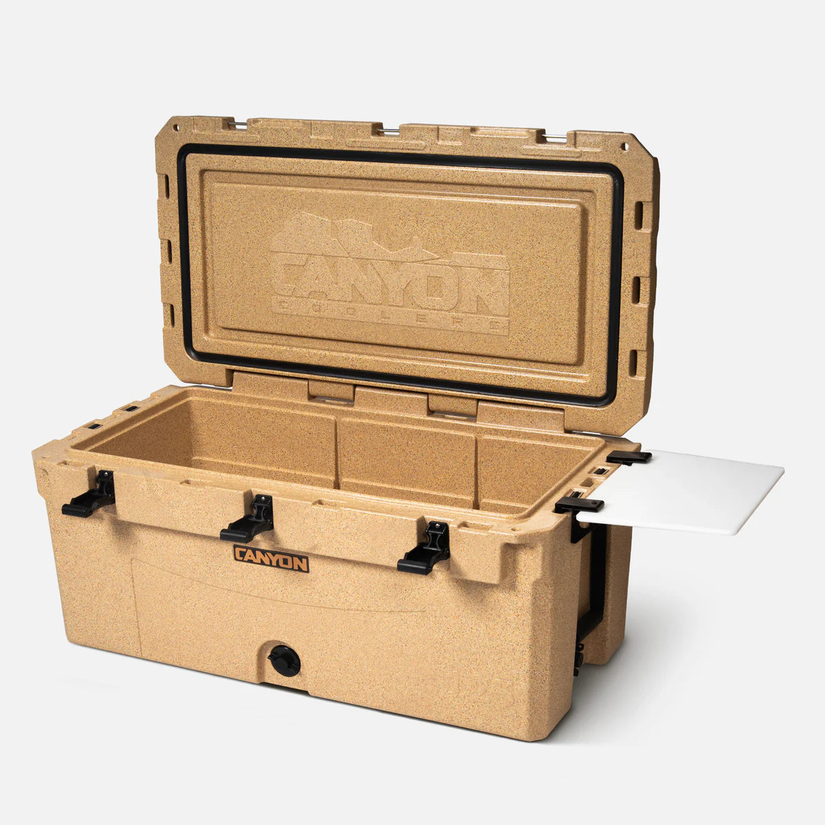 The tan Canyon Coolers Prospector 125 Raft Cooler features black latches, a drain plug, and a white cutting board partially extended—perfect as an expedition-sized hard-sided cooler for any adventure.