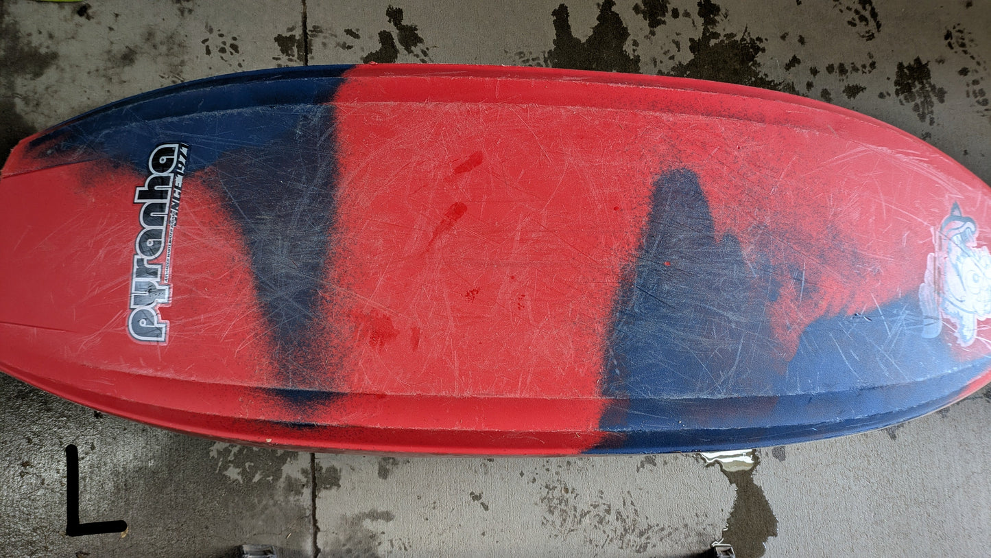 The Pyranha Demo Helectron kayak, in red and dark blue, is shown upside down on wet concrete with visible scuff marks and footprints—clear evidence of a well-used demo fleet model.