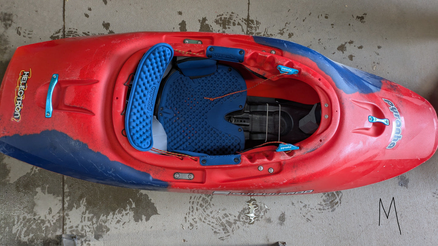 A Pyranha Demo Helectron used kayak from our demo fleet, featuring red and blue colors, padded blue seat inserts, and visible footrests, rests upside-down on a wet concrete surface.