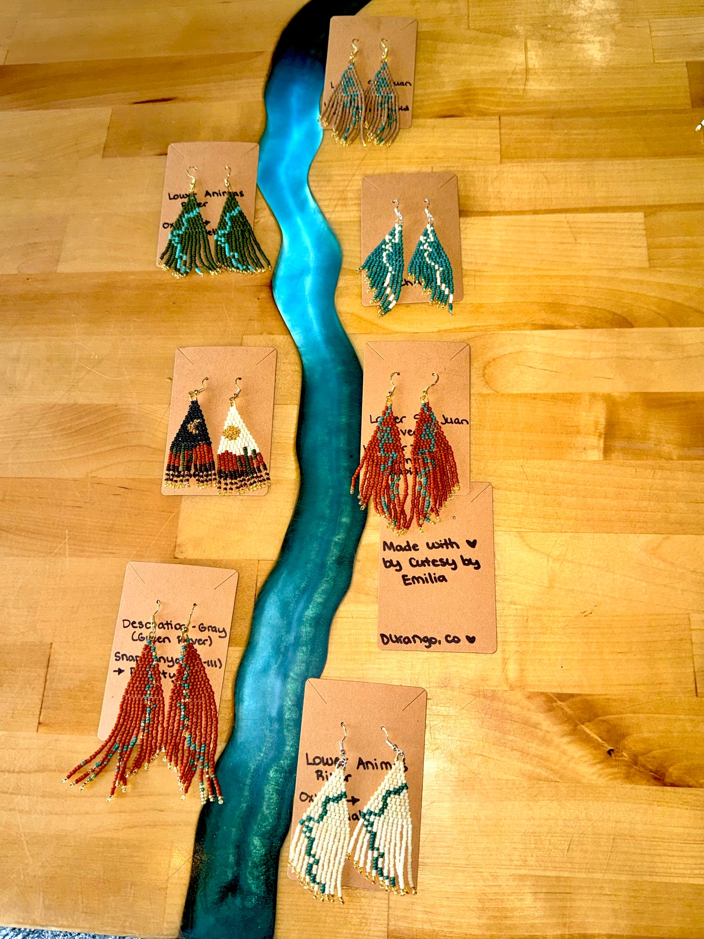 Seven pairs of 4CRS's Cutesy By Emilia Earrings, each with vibrant colors and unique designs like topographic views and desert flowers, are displayed on brown cards atop a wooden table featuring a striking blue resin river.