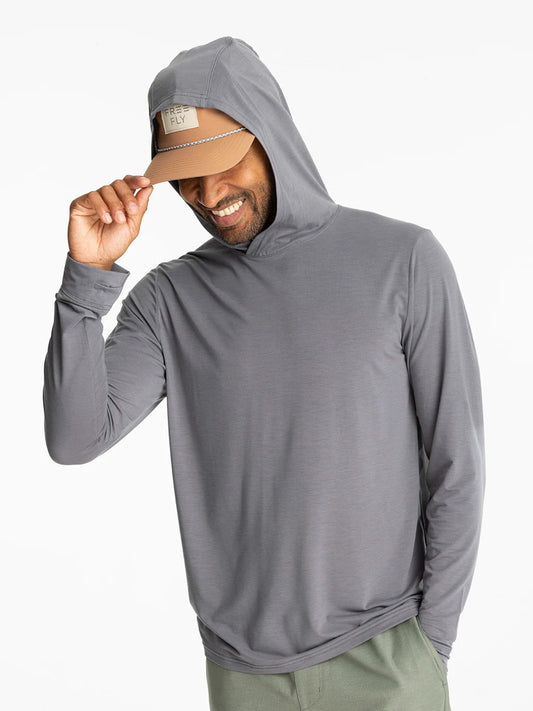 A man in a gray Free Fly Elevate Lightweight Hoodie M's with drirelease tech and a beige cap smiles, tilting his cap with one hand against a plain white background.