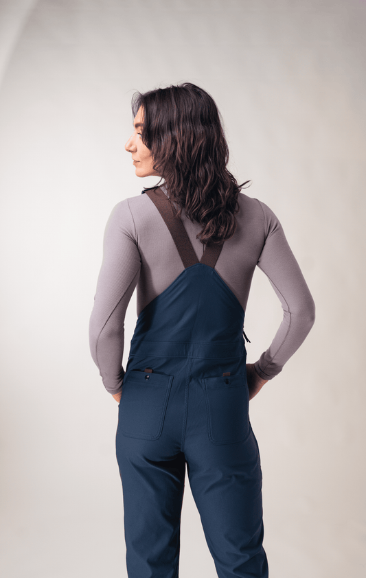 A person with long brown hair faces away, wearing the Allover Lookout Overalls 2.0 W's—navy blue with adjustable straps and functional pockets—paired with a gray long-sleeve shirt.