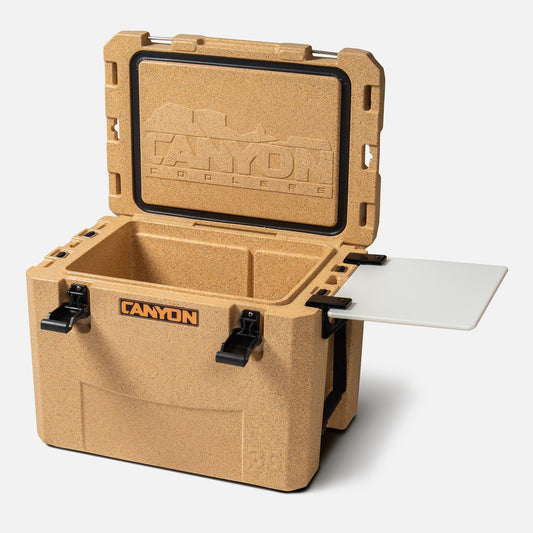 A tan Canyon Coolers ice chest with open lid, black latches, and a white Side Table / Divider made of HDPE attached to the side, plus space for an Outfitter V2 cooler divider.