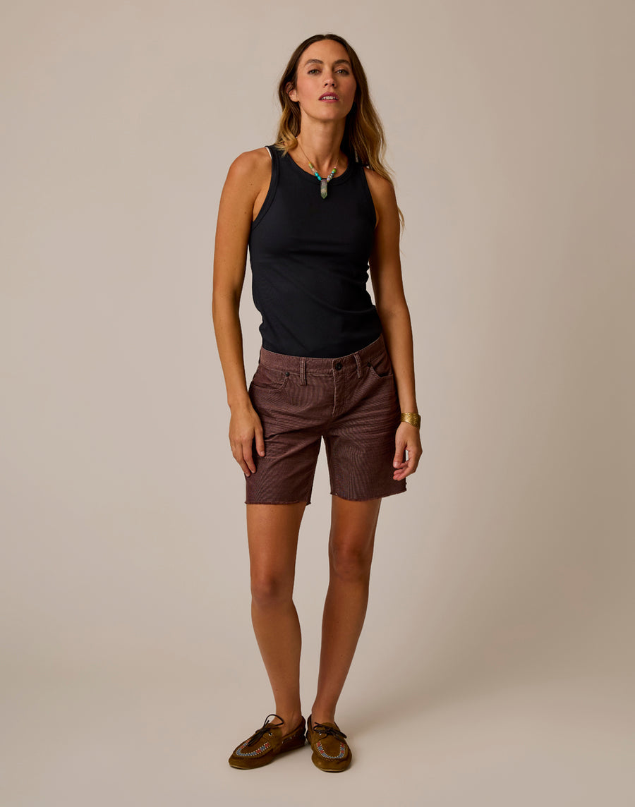 A woman stands against a plain background wearing a black sleeveless top, Carve Oahu 6" Shorts in maroon stretch cotton corduroy, brown shoes, and a green pendant necklace—ideal for refreshing your vacation style.