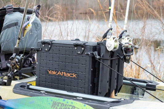 A YakAttack BlackPak Pro Kayak Fishing Crate with rod holders, fishing rods, and a tackle bag is mounted on a kayak by a lake, surrounded by dry grass.