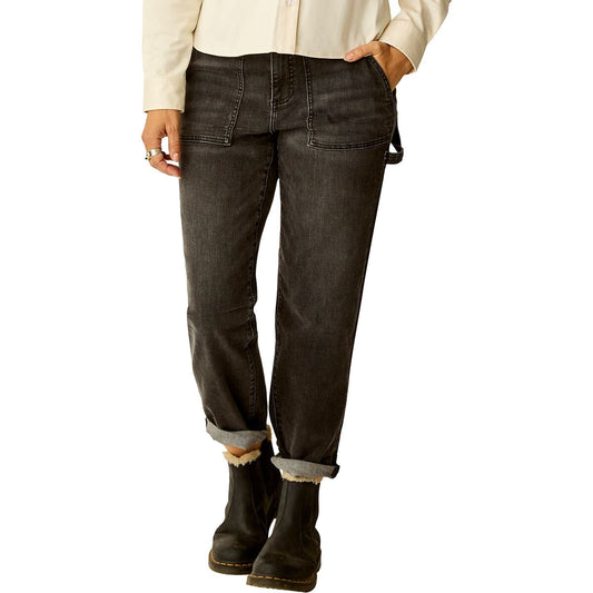 A person wearing Carve’s Corey Denim Pant in black, paired with black boots and a light-colored long sleeve shirt, stands with one hand in the front pocket.